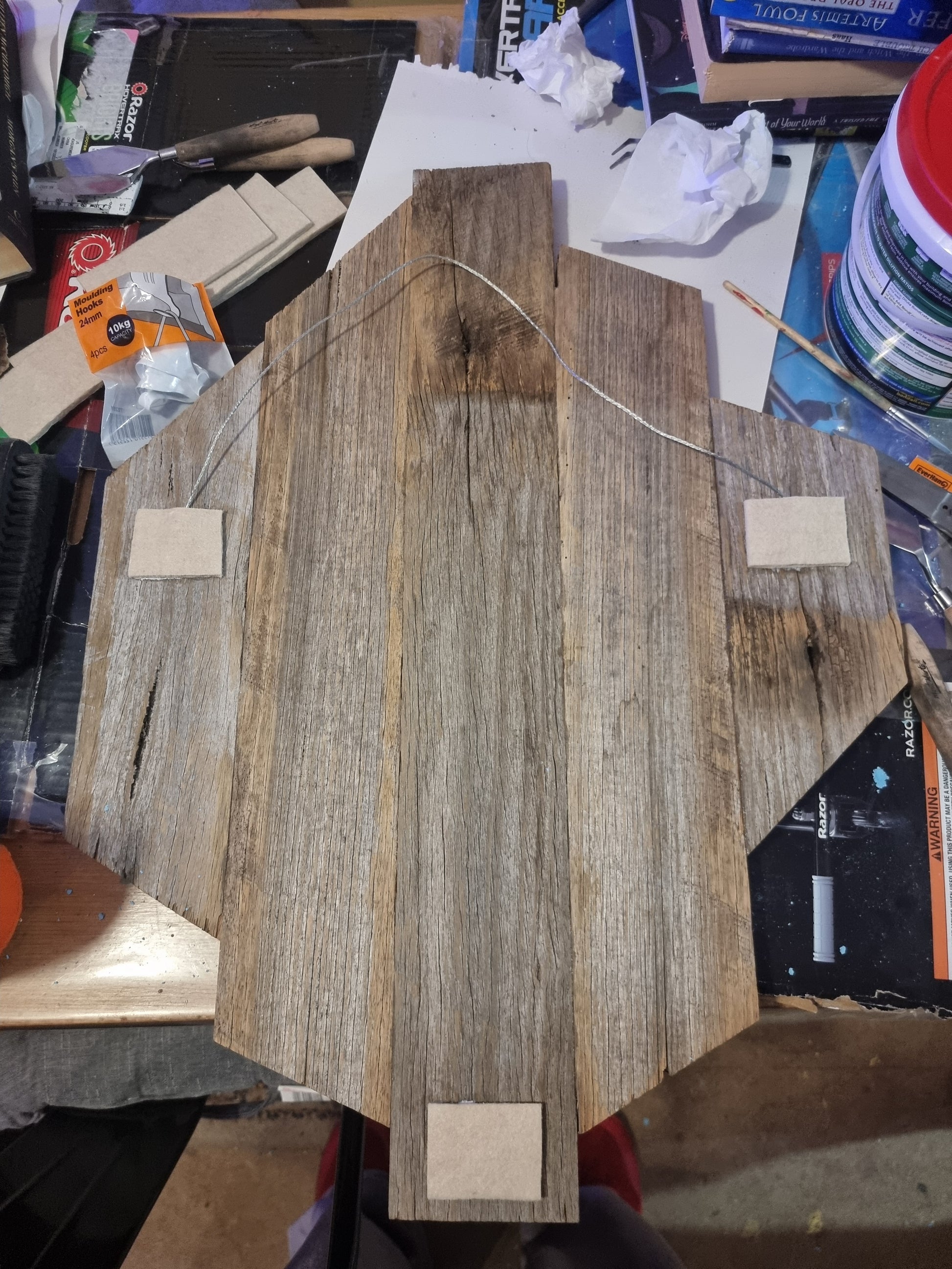 The back of a rustic wooden mosaic frame, showing installed hanging hardware — a metal wire secured between two points — and three felt pads placed at key contact points to protect the wall from scratches. The frame rests on a cluttered workbench with tools, packaging, and craft supplies nearby.