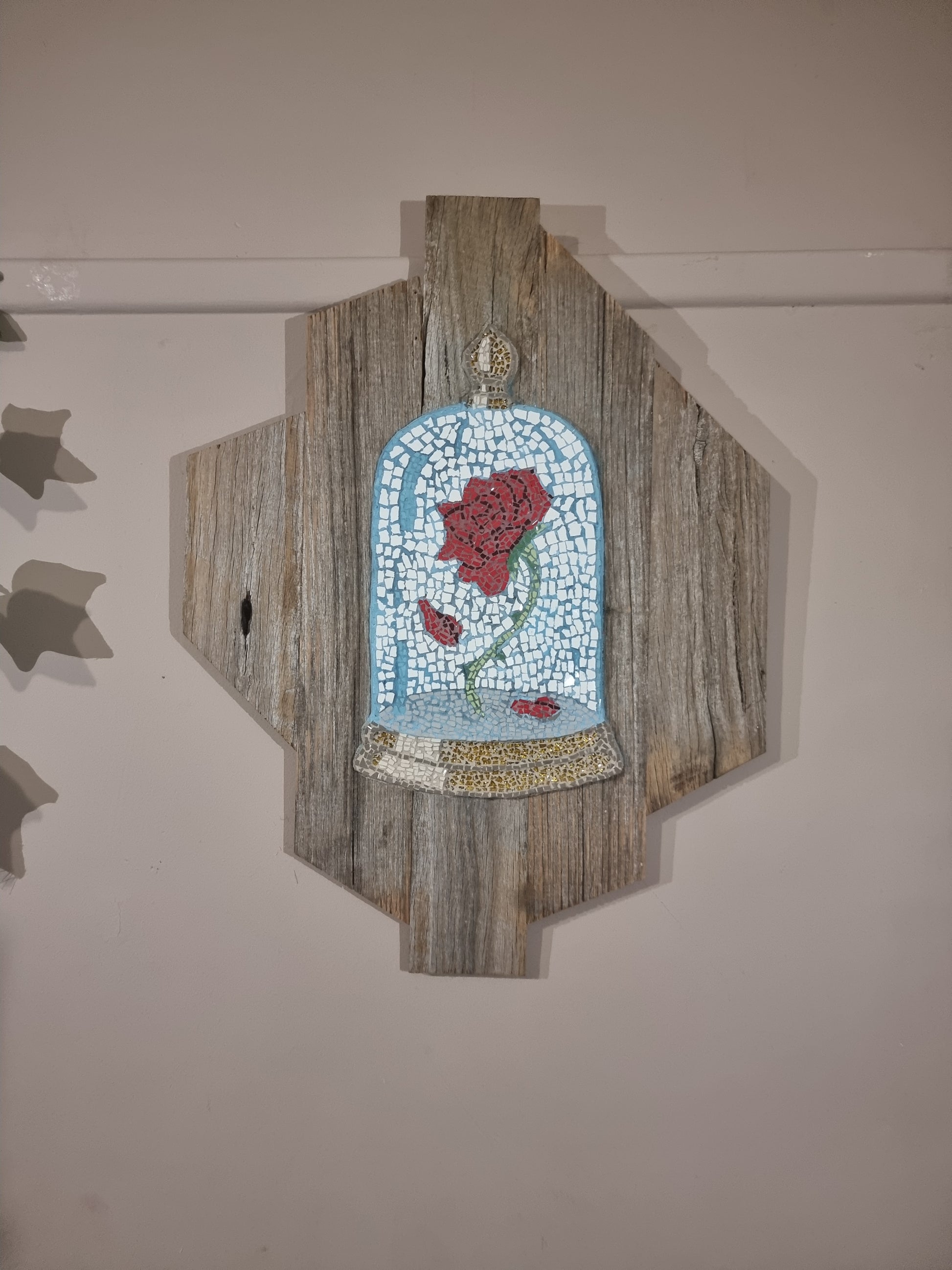 A mosaic artwork mounted on a rustic wooden panel, depicting a single red rose under a glass dome, inspired by "Beauty and the Beast." The rose is surrounded by falling petals and set against a pale blue background, with a detailed mosaic base resembling stone or gold. The artwork hangs on a light-colored wall with subtle shadows.