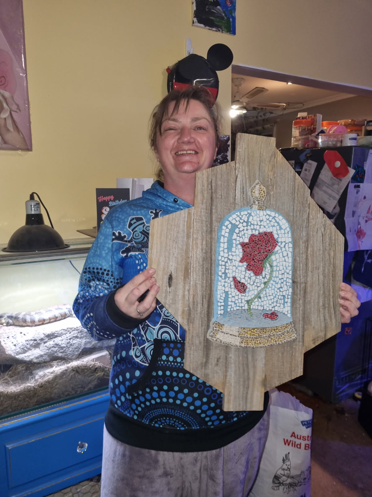 A smiling Merrin stands indoors holding a rustic wooden mosaic featuring a red rose under a glass dome, inspired by "Beauty and the Beast." She wears a blue patterned hoodie and grey pants. Behind her is a blue cabinet with a reptile enclosure on top and various household items in the background.