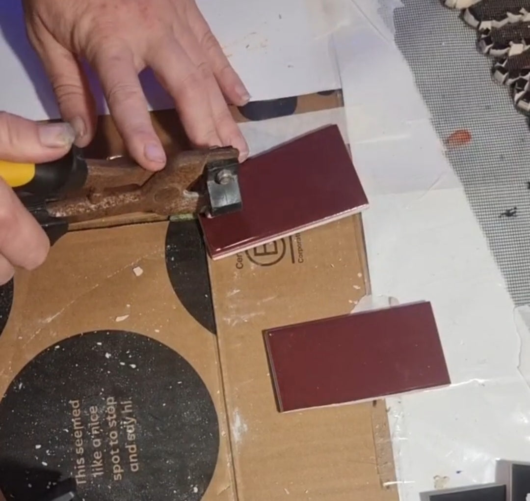 Load video: Video still showing maroon tiles being cut into strips and then carefully nipped into small pieces for mosaic use. Hands and tile-cutting tools are visible working precisely on a work surface.