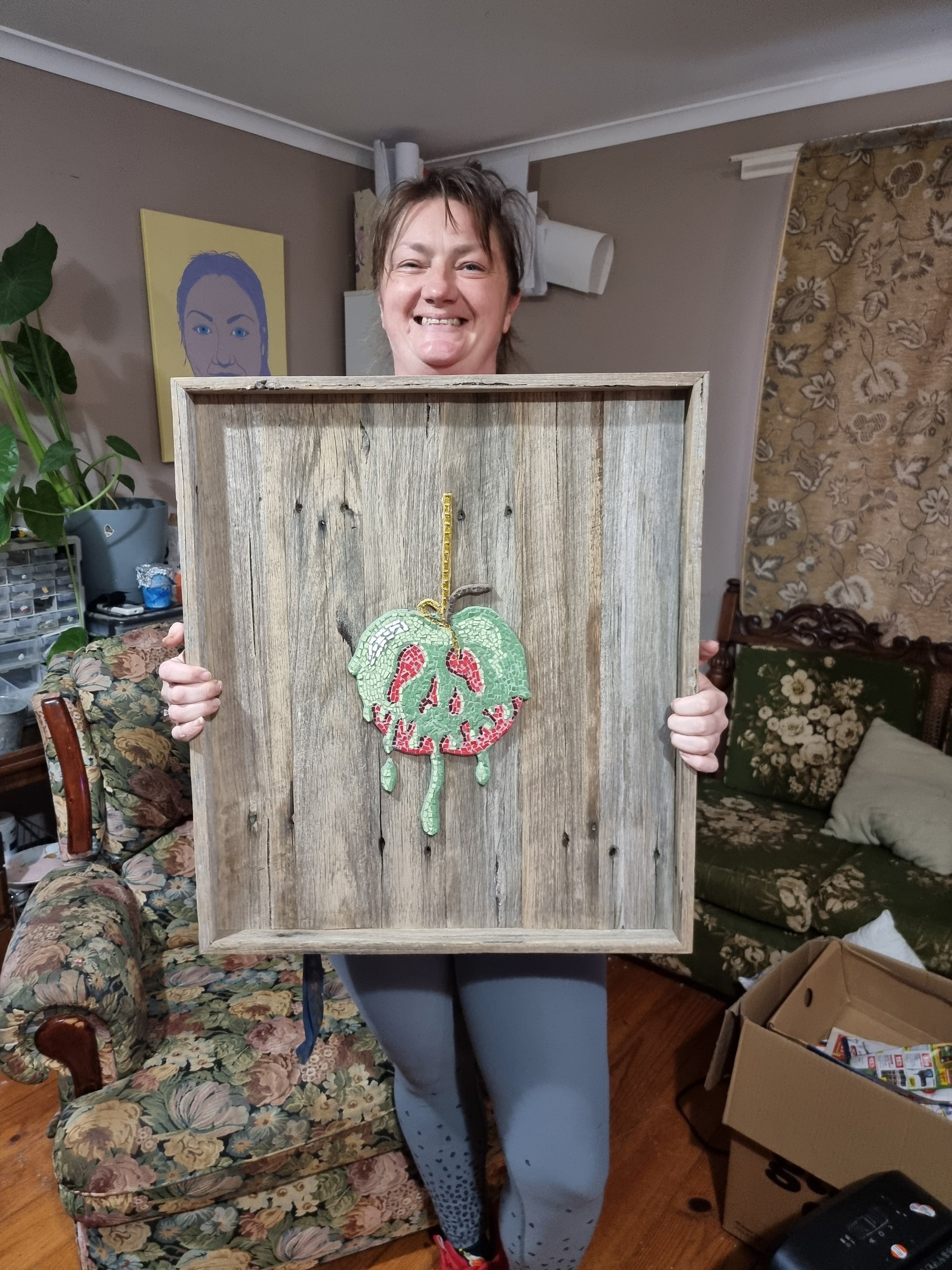 Artist holding the completed Poisoned Apple mosaic, mounted in a shadow box frame. The mosaic features rich, dark tones and detailed tiling. The piece is presented proudly in front of the artist’s studio.