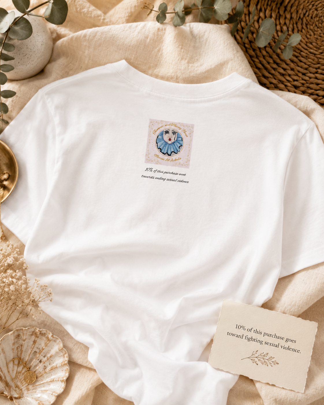 Back of the Willow t-shirt showing the Shimmer and Whimsy House logo and giving message, styled with soft linen, eucalyptus and a card reading “10% of this purchase goes toward fighting sexual violence.”