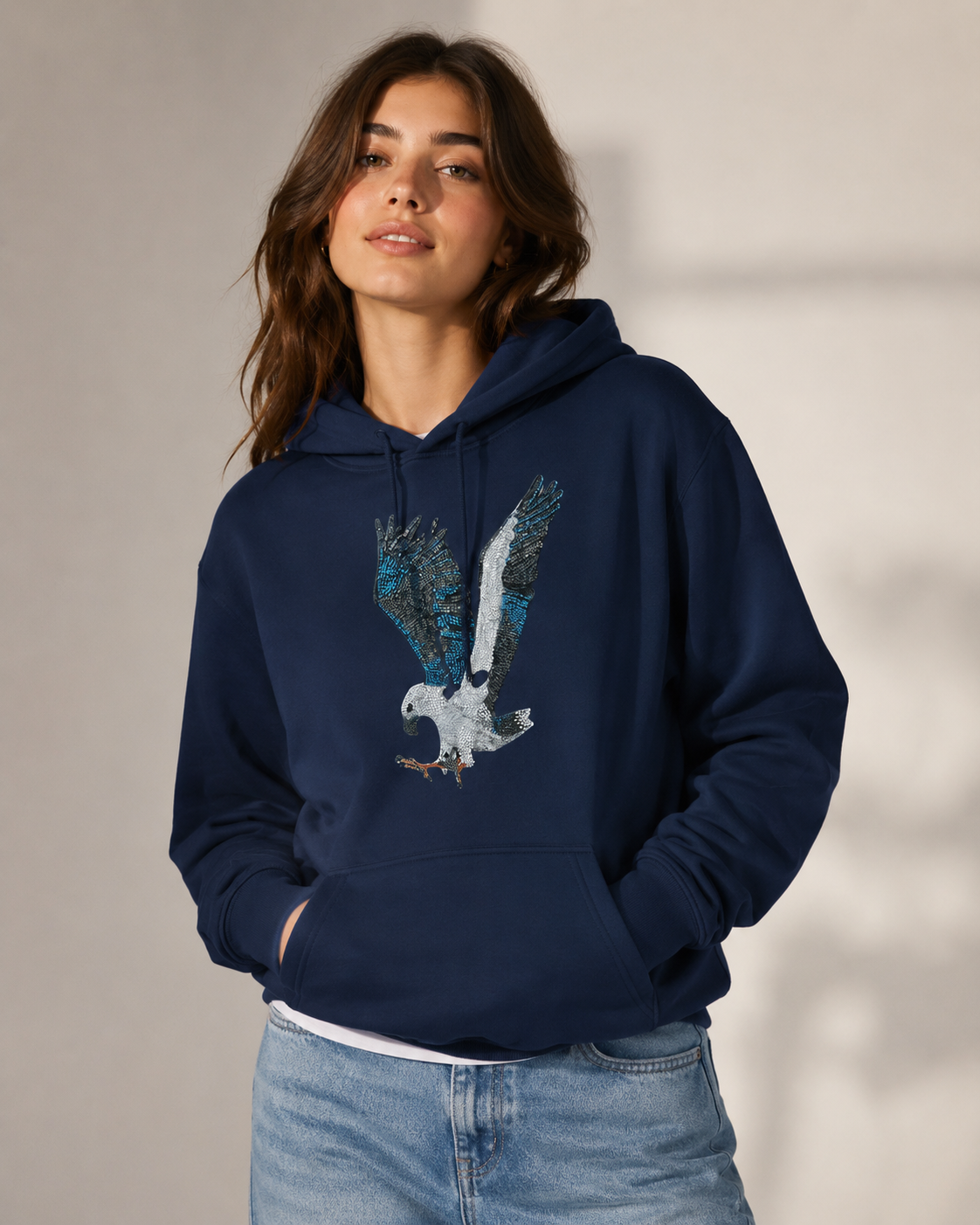 Fashion-style studio image of a model wearing the Willow mosaic hoodie with relaxed denim styling, natural makeup, soft lighting, and the White-bellied Sea Eagle artwork centred on the chest.