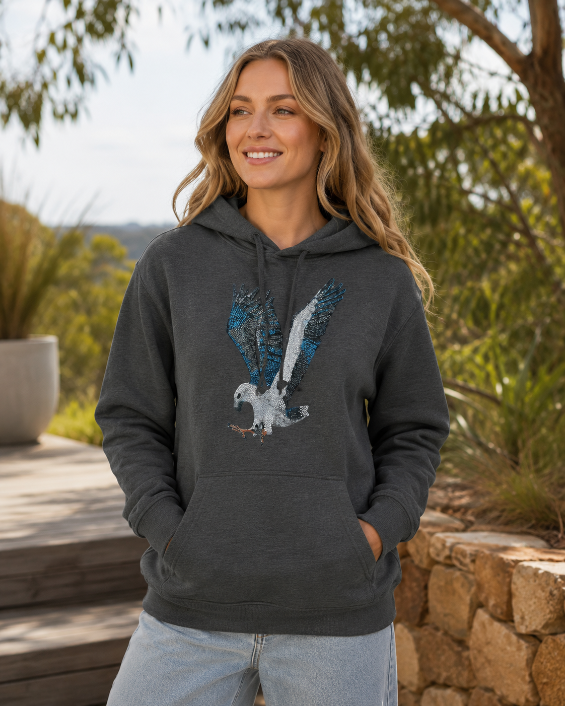 Model wearing the Willow White-bellied Sea Eagle mosaic hoodie outdoors near native Australian greenery, with soft natural light highlighting the cosy fit, dark hoodie fabric, and eagle artwork.