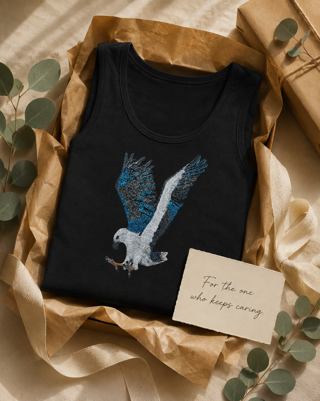 Gift-style image of the Willow tank top folded with recycled paper, soft ribbon, eucalyptus, and a handwritten card reading “For the one who keeps caring,” styled as a thoughtful wearable art gift inspired by the Willow white-bellied sea eagle mosaic story.