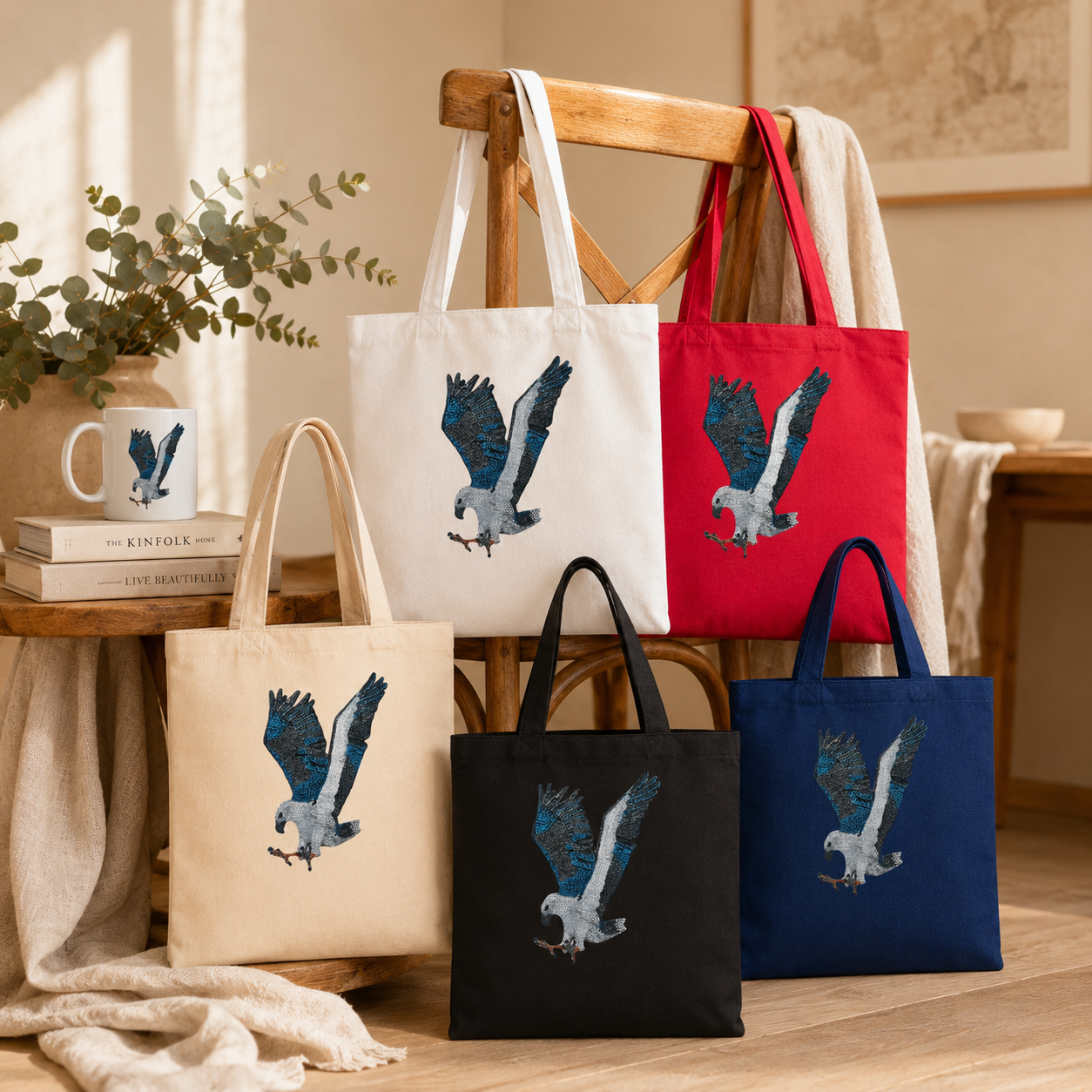 Full Willow tote bag colour range displayed in a warm lifestyle setting with timber furniture, soft linen, eucalyptus, and golden natural light, showing white, natural, black, navy, and red reusable cotton totes with the White-bellied Sea Eagle mosaic artwork clearly visible.