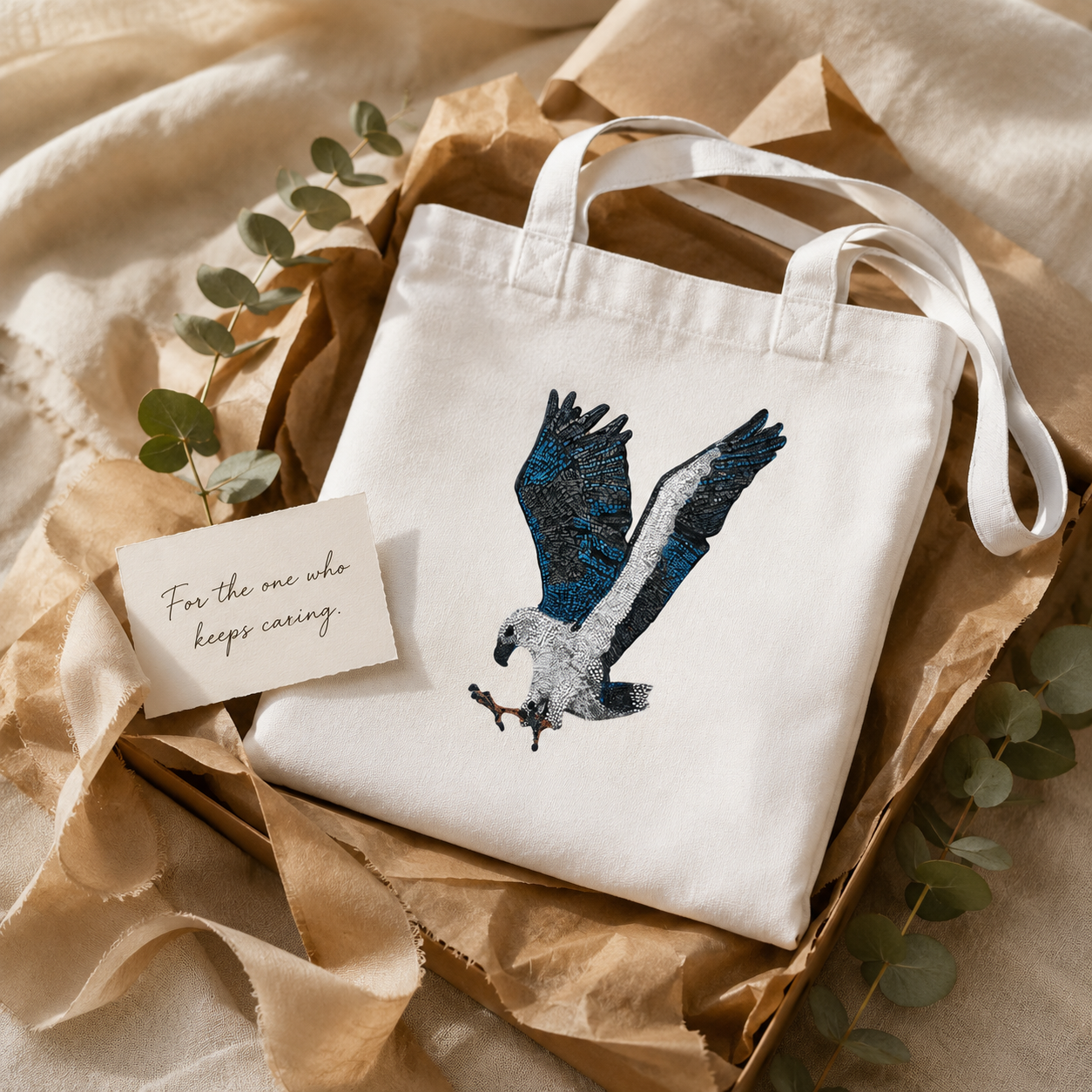 Gift-style Willow tote bag folded with recycled paper, soft ribbon, eucalyptus leaves, and a handwritten card reading “For the one who keeps caring,” showing the White-bellied Sea Eagle mosaic artwork as a thoughtful nature-inspired gift for art lovers, wildlife lovers, and caring souls.