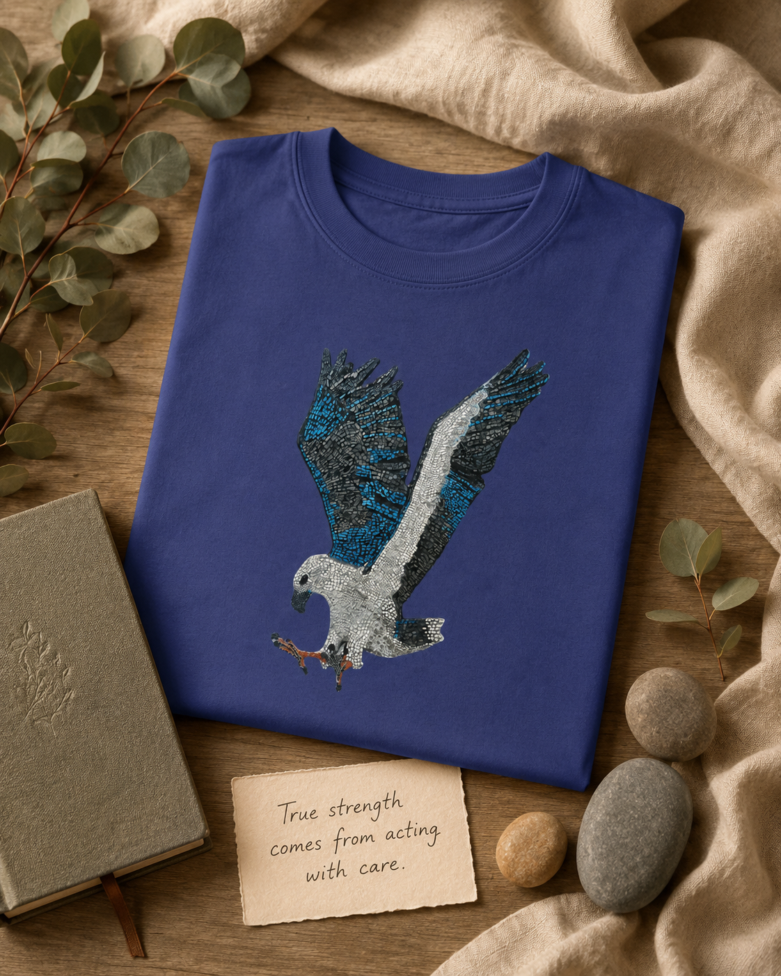 Folded Willow White-bellied Sea Eagle mosaic t-shirt styled with eucalyptus, river stones, soft linen, a journal and a handwritten quote card reading “True strength comes from acting with care.”